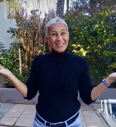 Person with short grey hair, wearing a black top, smiling and gesturing with open hands.